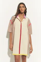 Vestido corto tipo túnica color crema con bloques de color beige en hombros y laterales. Presenta cuello camisero rosa, cierre frontal con línea roja y terminación con franja amarilla. Detalles de líneas turquesas y fucsias en hombros.