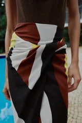 Falda midi de corte cruzado con diseño tipo pareo, presenta un nudo decorativo en la parte delantera y cierre de cremallera oculta en la espalda. El tejido cuenta con un estampado geométrico abstracto en tonos bordó, negro, amarillo y crema.