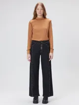 Jeans de corte ancho (wide leg) y tiro medio, confeccionados en denim de algodón negro con costuras contrastantes en color ocre. Presenta múltiples botones metálicos frontales y tachas metálicas aplicadas a lo largo de los laterales y la pretina.