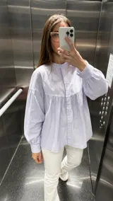 Camisa de corte amplio y diseño holgado, confeccionada en algodón. Presenta cuello mao, cierre frontal con botones, mangas largas abullonadas con puños abotonados y un detalle de frunces en la parte superior del pecho.