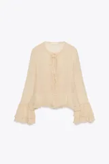 Camisa de viscosa con cuello redondo y escote en pico ajustable mediante lazadas. Presenta cierre frontal con botones, mangas largas con terminación evasé y detalle de volantes en el mismo tejido.