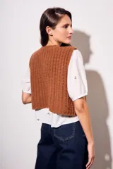 Chaleco tejido color beige con cuello redondo, sin mangas, con cierre frontal de cuatro botones dorados y dos bolsillos frontales.