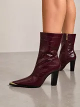 Bota de caña media en cuero vacuno con textura de reptil, color bordó. Presenta punta fina con puntera metálica dorada y taco cuadrado alto de color negro. Incluye cierre lateral interno.