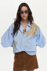 Camisa de algodón a rayas celestes y blancas, con mangas abullonadas y gran estampado floral en el frente.