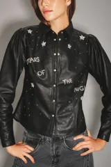 Camisa de cuero ovino color negro, con mangas abullonadas, cuello clásico y cierre frontal con botones metálicos. Presenta detalles bordados con pedrería en forma de estrellas y la frase 'Stars of the night'.