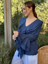 Camisa de lino azul marino con volados en cascada a lo largo del frente y en los puños de las mangas largas. Presenta un escote en V con un botón de nácar visible.