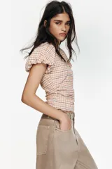 Blusa de manga corta abullonada con cuello redondo, confeccionada en tela a cuadros. Presenta detalle de frunces en los laterales y cierre lateral con cremallera oculta.