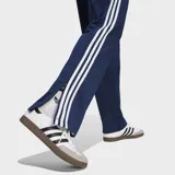 Pantalón deportivo Adidas Firebird Drop Needle de color azul marino, con las clásicas tres tiras blancas en los laterales y el logo del Trifolio bordado. Presenta un diseño de rayas verticales sutiles en relieve.