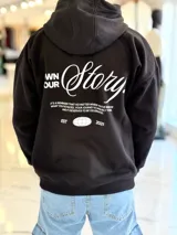 Canguro oversize negro con capucha y bolsillo tipo canguro. Presenta un gran estampado en la espalda con la inscripción "Own Your Story", texto descriptivo y un gráfico de globo terráqueo junto al año "2021".