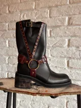 Bota alta de cuero negro con diseño estilo western, tiras en tono bordó con tachas metálicas, hebilla superior y arandela dorada en el tobillo. Cuenta con taco ancho de efecto madera.