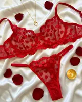 Conjunto de lencería de dos piezas rojo confeccionado en tul con diseño de corazones bordados. Incluye corpiño con aro y tirantes regulables, y bombacha colaless regulable con detalle de letras de strass.