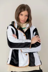 Campera de textura suave y afelpada con diseño de rayas verticales en blanco y negro. Presenta un corte recto, cuello con solapas y cierre frontal con botones. Los bordes están terminados con un ribete en color tostado que aporta contraste y estructura a la prenda.