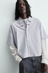 Camisa de corte relajado con diseño de rayas verticales en tonos azul y blanco. Presenta cuello solapa, cierre frontal de botones y un detalle de texto bordado en color rojo en el pecho. Cuenta con un diseño de manga larga con efecto doble, combinando la manga corta de la camisa con una manga larga de tejido tipo waffle en color crudo.