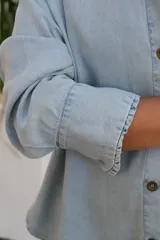 Camisa de jean en tono celeste claro, de corte holgado y diseño oversize. Presenta cuello clásico, cierre frontal con botones y mangas largas con puños con detalle de volado sutil.