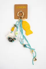 Charm para bolso con argolla dorada, cordones de hilo celeste y colgantes variados que incluyen cuentas de cerámica, conchas marinas, abalorios de cristal y un dije grande en forma de pez de color amarillo.