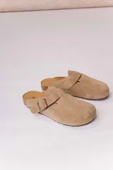 Zueco de gamuza color tostado, con diseño cerrado en la parte delantera, tira ajustable con hebilla metálica en el empeine, plantilla anatómica y suela tipo corcho.