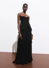 Vestido maxi largo negro de viscosa, palabra de honor y sin mangas. Presenta diseño evasé con detalle de volantes escalonados y cierre de cremallera lateral.