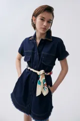 Vestido corto de denim azul oscuro, con cuello camisero, mangas cortas con dobladillo y cierre frontal con botones. Tiene bolsillos en el pecho y laterales, y cinturón de pañuelo estampado.