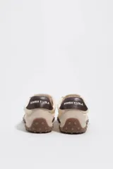 Zapatillas deportivas de color beige y crudo, confeccionadas en serraje vacuno con inserciones de tela, cordones marrones y suela de caucho con tacos. Cuenta con detalle de piel marrón en la parte trasera con logo.
