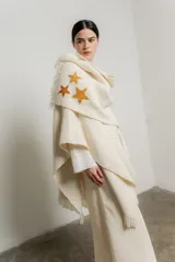 Ruana de tejido de punto color crudo, con flecos en el ruedo y apliques de estrellas bordadas a mano en tonos dorados y ocres.