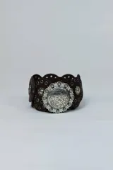 Cinto ancho de cuero marrón con detalles troquelados en los bordes, ojetes metálicos y una gran hebilla redonda plateada con grabados ornamentales.