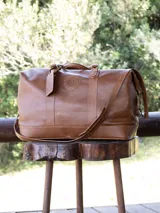 Bolso de viaje marrón de cuero liso con correa al hombro ajustable y extraíble. Tiene manijas superiores reforzadas, cierre superior con cremallera y etiqueta de cuero colgante.