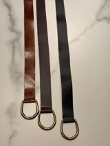 Cinto de cuero flexible y envolvente con diseño minimalista, que se anuda mediante una hebilla metálica en forma de D. El extremo presenta el logo de la marca grabado.
