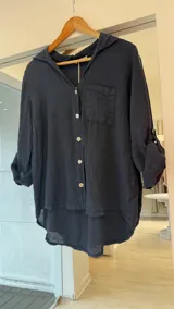 Camisa azul marino de lino con cuello camisero, cierre frontal con botones claros y un bolsillo aplicado en el pecho. Cuenta con mangas largas remangables y diseño de corte asimétrico, más larga en la parte trasera.