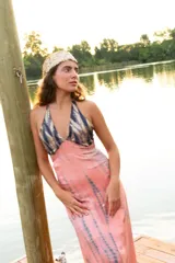 Vestido largo con estampado tie-dye en tonos violeta y fucsia, escote en V con detalles bordados dorados, cut-outs laterales bajo el busto y tirantes finos que se cruzan y atan en la espalda.