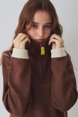 Buzo de tejido de punto color marrón con cuello alto y cierre frontal parcial. Presenta rayas verticales finas en color claro, puños y ruedo en tejido acanalado color crema, y detalles en color verde lima en el tirador del cierre, el cordón de ajuste del ruedo y el parche bordado en el pecho.
