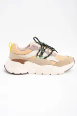 Zapatilla urbana de diseño chunky, con paneles combinados en tonos beige, arena y gris. Presenta cordones jaspeados en azul y amarillo, tirador trasero amarillo y suela de plataforma dentada en color crema.