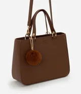 Cartera estilo satchel de tamaño mediano, confeccionada en poliuretano color marrón con textura tipo cuero. Cuenta con asas de mano reforzadas, correa larga desmontable, cierre superior de cremallera y un colgante decorativo de pompón de pelo sintético.