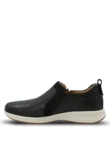 Zapatilla tipo slip-on Hush Puppies de cuero negro con cierre lateral decorativo, diseño de perfil clásico y suela de goma blanca con base antideslizante.
