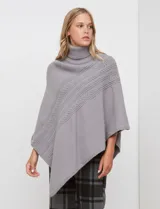 Poncho tejido color crema, con cuello polera y diseño trenzado diagonal en el frente. Presenta un corte asimétrico en el ruedo.