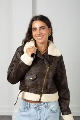 Chaqueta de estilo biker confeccionada en eco cuero con efecto gastado, presenta cuello y puños con forro de corderito sintético. Incluye cierre frontal cruzado, bolsillo pequeño con solapa y cinturón ajustable en la cintura.