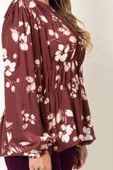 Blusa color marrón con estampado de flores blancas, de manga larga abullonada con puño elástico y cuello redondo con detalle de frunces horizontales.