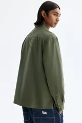 Camisa cargo color verde militar de corte regular, confeccionada en tejido ripstop de algodón. Cuenta con cuello inglés, cierre frontal de botones, bolsillos con solapa en el pecho y mangas largas con puños abotonados.