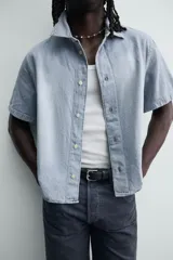 Camisa de corte boxy fit confeccionada en denim ligero de algodón. Presenta cuello solapa, manga corta y cierre frontal con botones. Diseño con efecto lavado.