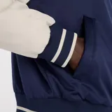 Campera estilo varsity con cuerpo azul marino y mangas en color beige. Presenta cierre frontal con botones a presión, cuello, puños y ruedo acanalados con rayas decorativas, y logo de la marca bordado en el pecho.
