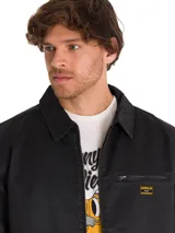 Chaqueta de estilo chore en color negro, confeccionada en sarga encerada. Presenta cierre frontal de cremallera metálica, cuello clásico, bolsillo en el pecho con cierre y etiqueta de la marca Caterpillar.