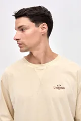 Remera de manga larga color beige, confeccionada en algodón lavado con textura suave. Presenta cuello redondo con detalle de costura en V, corte clásico y estampado pequeño en el pecho con la palabra Uniform.