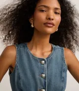 Chaleco de jean azul con cuello redondo y cierre frontal de botones metalizados con relieve. Presenta recortes verticales que definen la silueta y un bajo acabado en pico. Confeccionado en tejido de algodón denim.