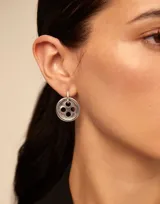 Par de aros asimétricos plateados. Uno es un aro con dije colgante en forma de aguja de coser. El otro es un aro con dije colgante en forma de botón con cuatro agujeros.