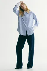 Camisa oversized de popelina de algodón con diseño a rayas verticales en tonos azul y blanco. Presenta cuello clásico y puños anchos en color blanco sólido, cierre frontal con botones, hombros caídos y bajo redondeado con aberturas laterales.