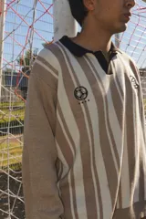 Buzo de punto tejido en jacquard con diseño de rayas verticales en tonos beige y blanco. Presenta cuello polo en color negro, puños y ruedo acanalados en blanco, y bordados alusivos al mundial en el pecho.