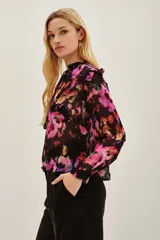 Camisa de viscosa con estampado floral en tonos fucsia, violeta y naranja sobre fondo negro. Presenta cuello con lazo ajustable y mangas largas con puños elásticos.