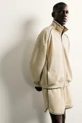 Bermuda de corte relajado confeccionada en tejido de algodón, color beige con vivos en contraste color verde oscuro. Cuenta con cintura elástica ajustable con cordón, bolsillos frontales y bolsillos traseros de plastrón.