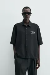 Camisa de corte relajado confeccionada en tejido fluido con mezcla de viscosa, color negro. Presenta cuello solapa, manga corta, cierre frontal con botones y un bordado a contraste en el pecho.