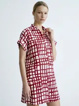 Vestido camisero de silueta relajada, con cuello clásico, cierre frontal de botones y mangas cortas con dobladillo. Presenta un estampado de cuadrícula irregular en color rojo sobre fondo blanco.