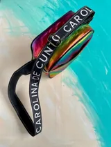 Bandolera Kaira de la línea Ottenta de Carolina De Cunto, confeccionada en material metalizado con acabado multicolor y efecto tornasolado. Cuenta con un bolsillo frontal con broche imantado, cierre superior y una correa larga y ajustable de tela negra con el nombre de la marca en letras blancas.